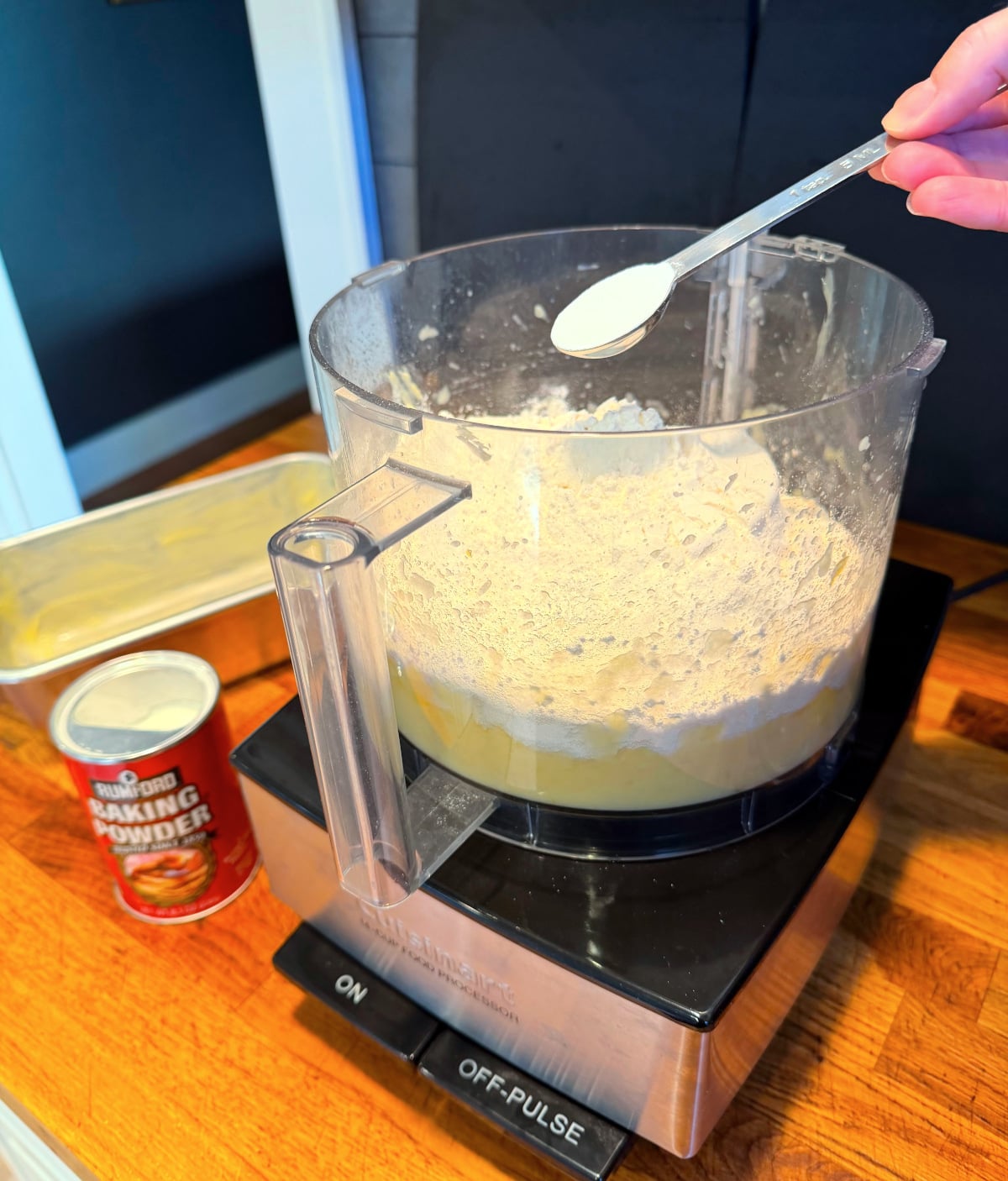 White powder being tipped from a steel measuring spoon into a food processor filled with flour, butter, and pureed apple next to a can of baking powder.