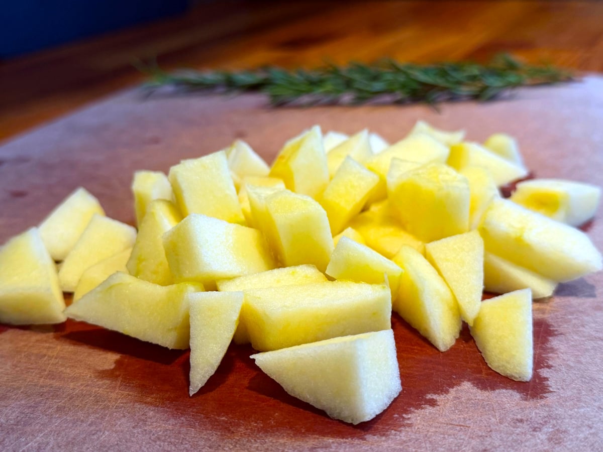 Chopped apples on a wooden cutting board in front of a couple sprigs of rosemary.