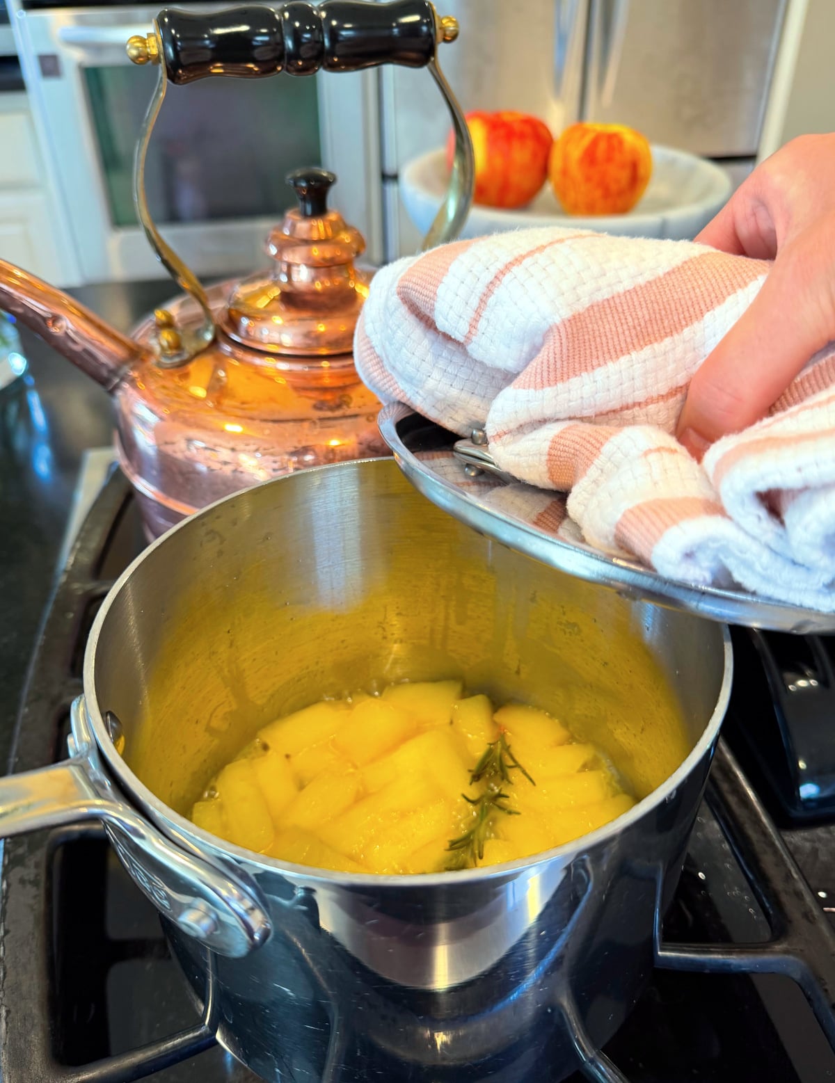 Lid being lifted using a pink and white striped towel from a small steel saucepan with apples and rosemary cooking on the stove.