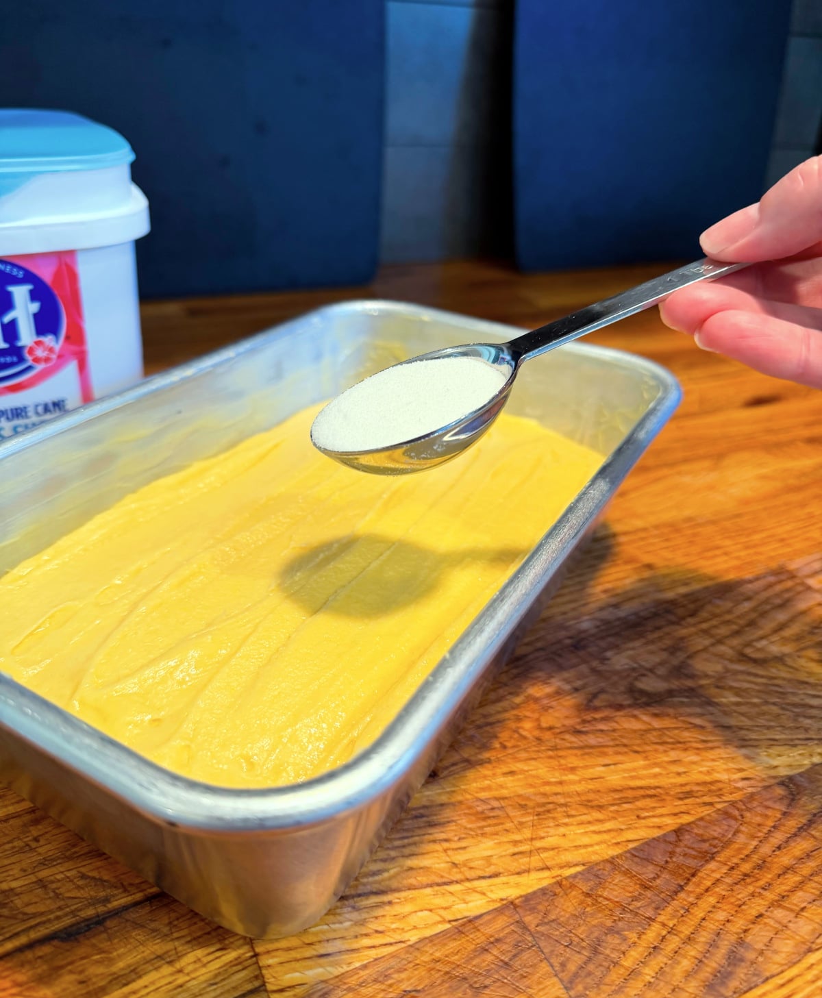 Sugar being sprinkled from a steel measuring spoon over golden yellow cake batter in a metal loaf pan.