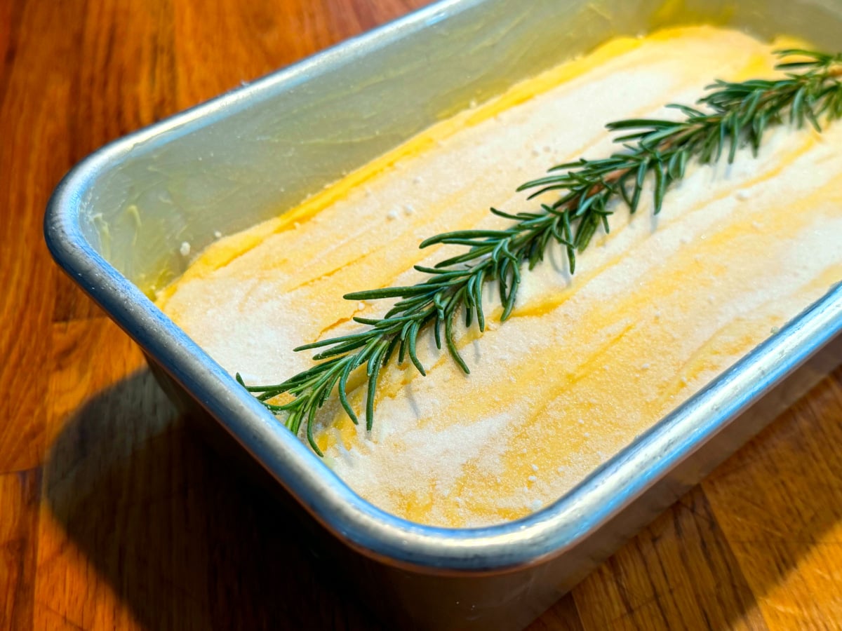 Golden yellow cake batter sprinkled with sugar and decorated with rosemary sprig in a metal loaf pan.