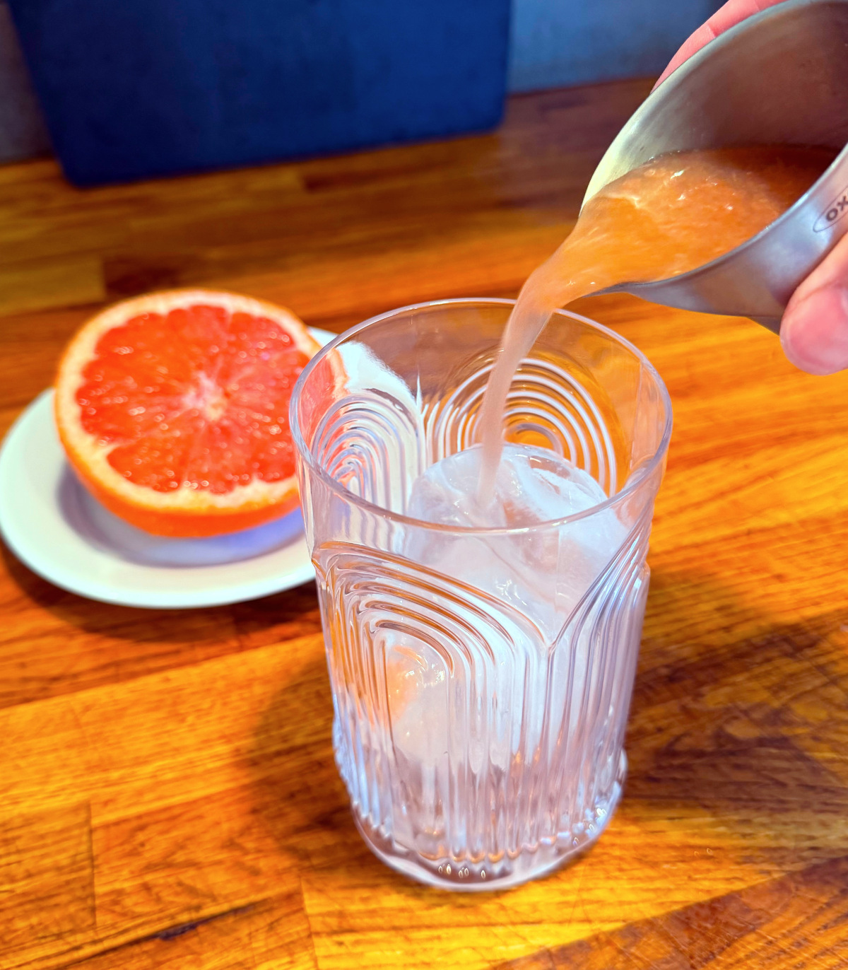 Pale pink liquid being poured into a tall etched glass full of ice next to half a grapefruit sitting on a small white plate.