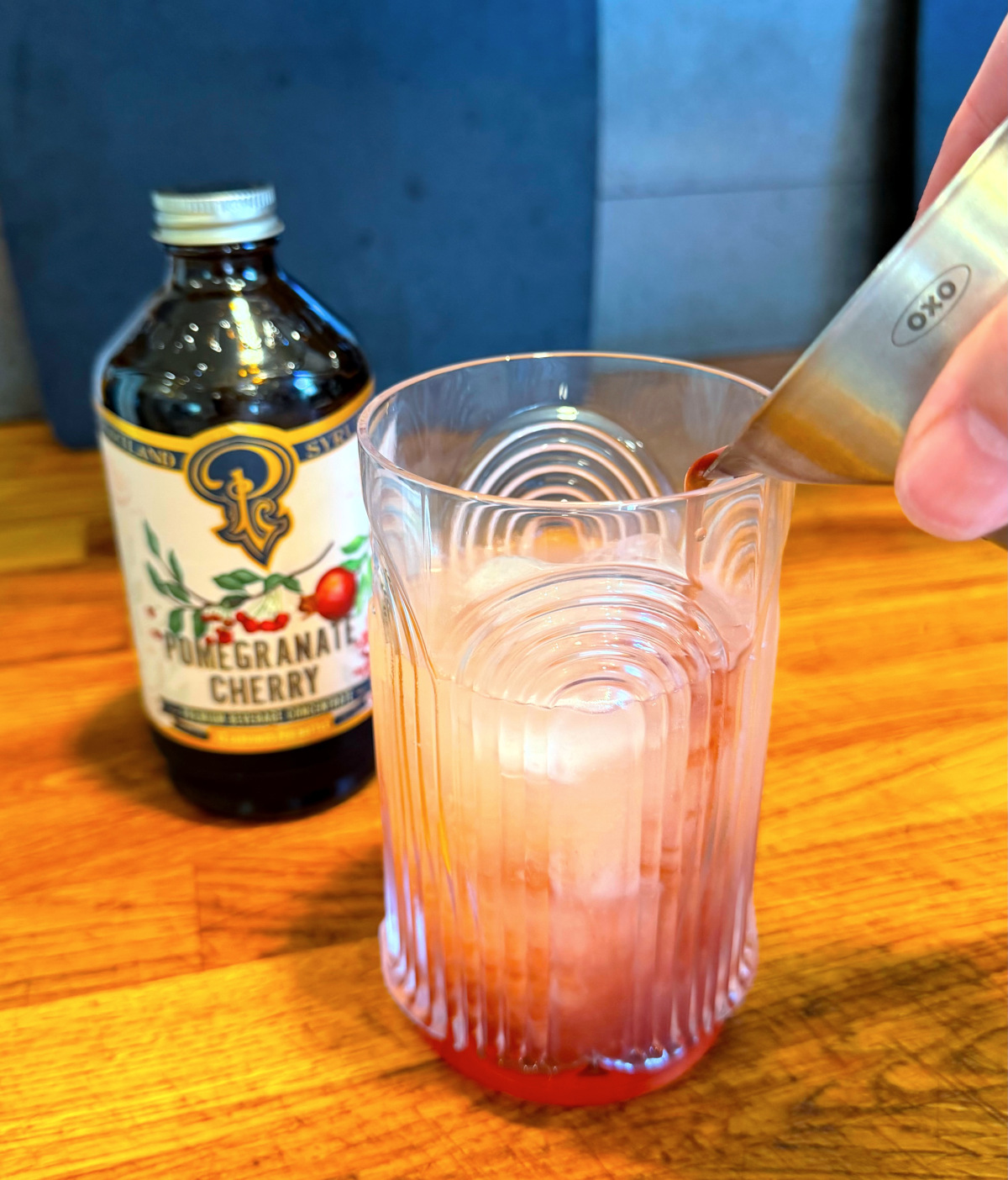 Dark red liquid being poured from a steel measuring jigger into a tall etched glass containing pale pink liquid next to a bottle of grenadine.
