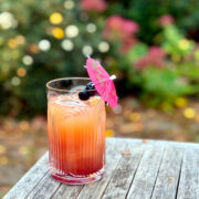Tequila sunset served in a tall etched glass garnished with two maraschino cherries skewered on a pink drink umbrella and sitting on an outdoor wooden table in front of some shrubs.