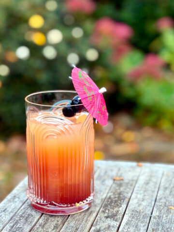 Tequila sunset served in a tall etched glass garnished with two maraschino cherries skewered on a pink drink umbrella and sitting on an outdoor wooden table in front of some shrubs.