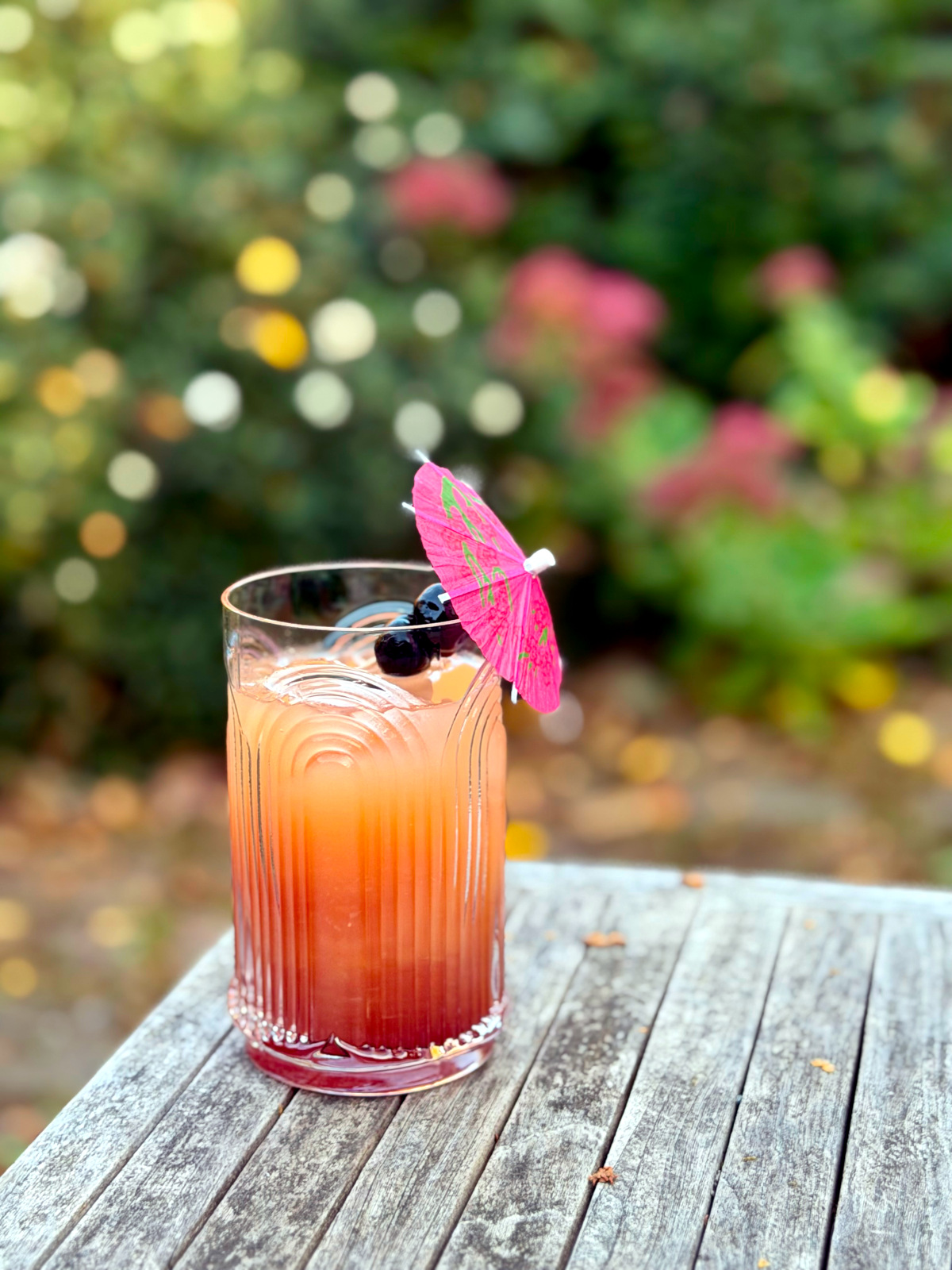 Tequila sunset served in a tall etched glass garnished with two maraschino cherries skewered on a pink drink umbrella and sitting on an outdoor wooden table in front of some shrubs.