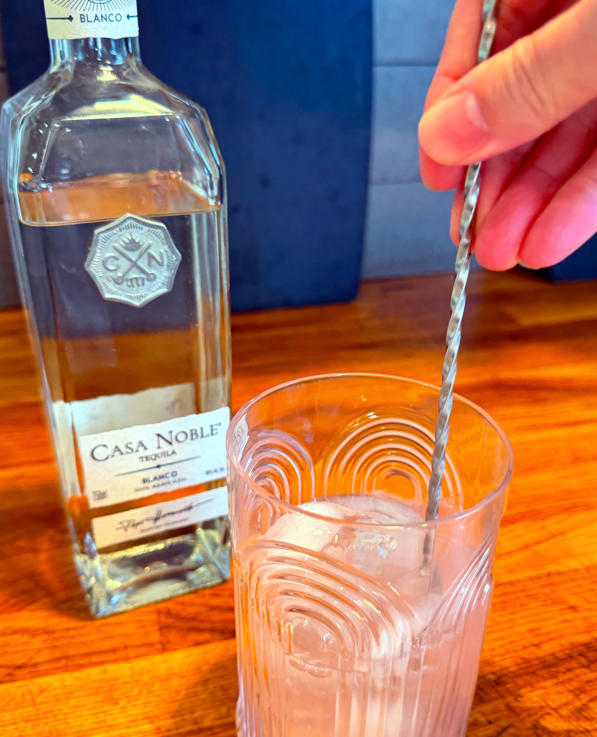 Pale pink liquid being stirred in a tall etched glass with a steel bar spoon next to a bottle of tequila.